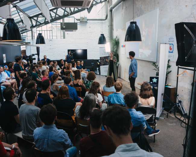 A large audience is seated in a modern venue as two speakers engage in a presentation on stage.
