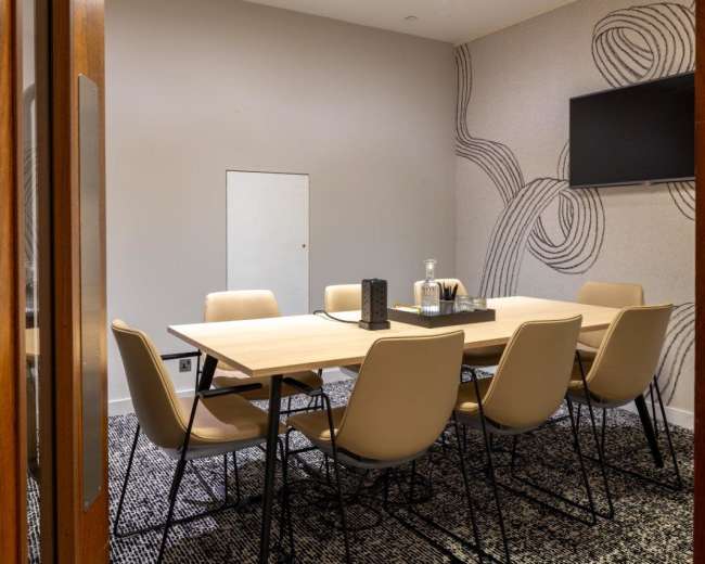 A modern conference room features a large wooden table surrounded by six beige chairs, with a wall-mounted television and decorative wallpaper.