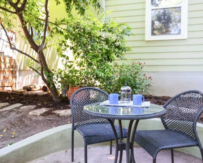 A small patio area features a round glass table with two chairs, surrounded by greenery and a light green wall.