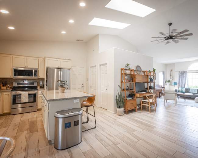 A bright, open-concept living area features a kitchen with light-colored cabinetry, a dining space with bar stools, and a cozy sitting area with large windows.