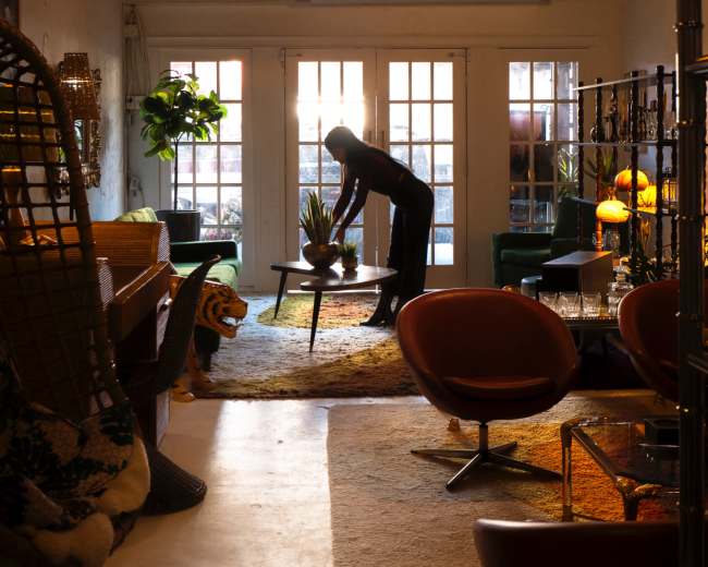 A person is arranging a plant on a table in a brightly lit room filled with various furniture and decor items.