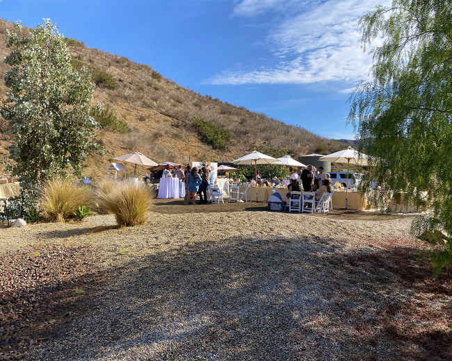 A gathering is taking place outdoors on a gravel area, with tables under umbrellas and a hillside in the background.