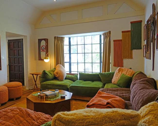 A cozy living room with green and orange furniture, a wooden table, and a large window letting in natural light.