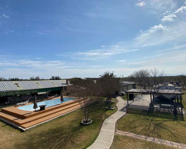 A backyard with a swimming pool surrounded by wooden decking, a paved pathway, and several buildings under a clear blue sky.
