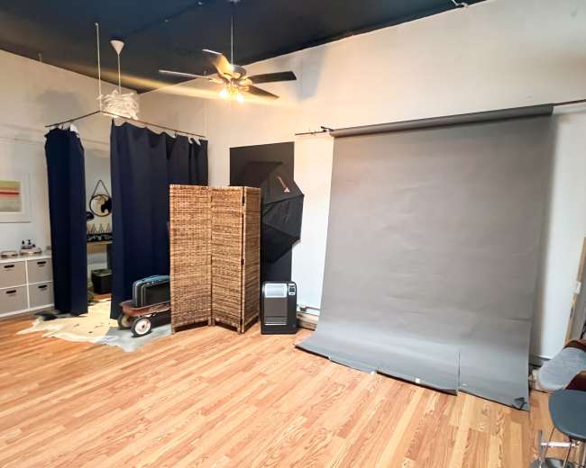 A spacious room with wooden flooring, featuring a gray backdrop, a screen divider, a fan, and various pieces of furniture.