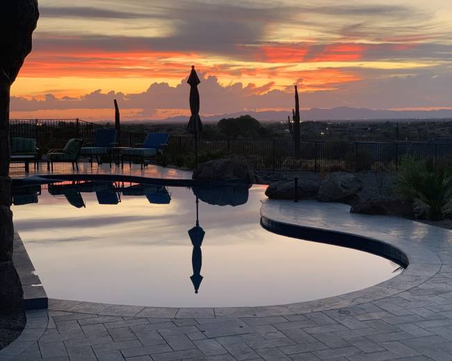 The scene features a swimming pool reflecting the vibrant colors of a sunset over distant mountains, surrounded by outdoor furniture and rocky landscaping.