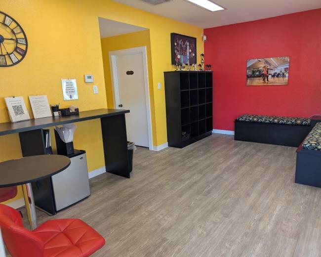 The room features yellow and red walls, a black shelving unit, a small table with a chair, and seating along the wall with decorative cushions.
