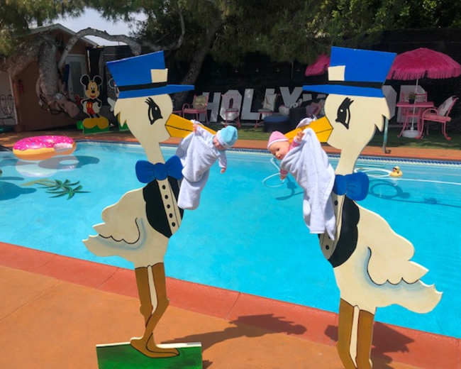 Two stork decorations holding newborn dolls stand beside a swimming pool surrounded by colorful inflatable toys.