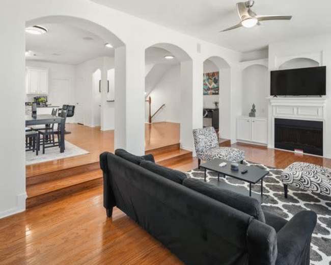 The image shows a modern living room with a black sofa, wood flooring, and a ceiling fan, adjacent to a dining area and staircase.