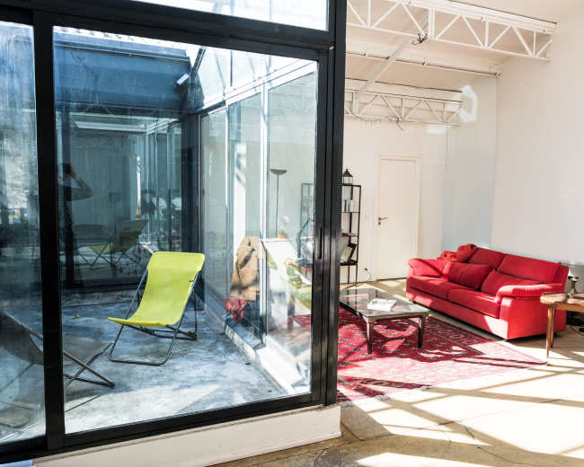 The image shows a modern, bright room with a red sofa, a yellow lounge chair, and large glass walls that provide views of an adjacent sunroom.