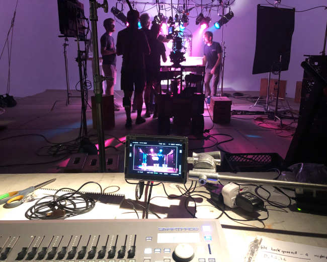 A film set with crew members gathered around a lighting setup, viewed from a controlled desk with equipment and a monitor displaying the scene.