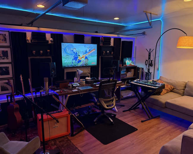 The image shows a music studio featuring a desk with a computer and audio equipment, surrounded by guitars, monitors, and illuminated by blue LED lights.
