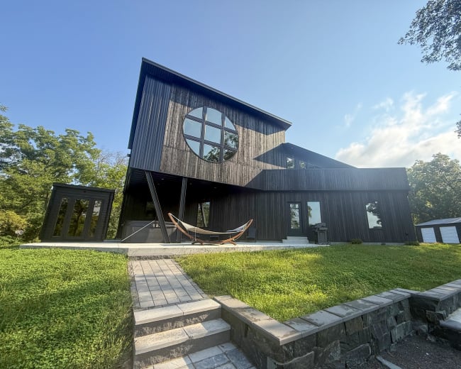 The image shows a modern, black wooden house with a large circular window, a stone pathway, and a hammock in the front yard surrounded by greenery.