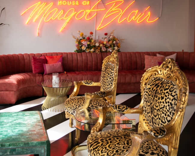 A stylish lounge area featuring leopard print chairs, a green coffee table, and neon signage that reads "House of Margot Blair."