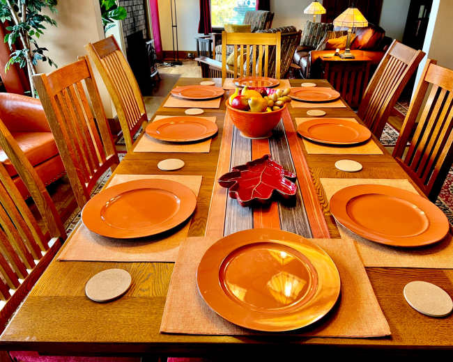 A wooden dining table is set with orange plates and placemats, featuring a centerpiece of fruit in a decorative bowl.