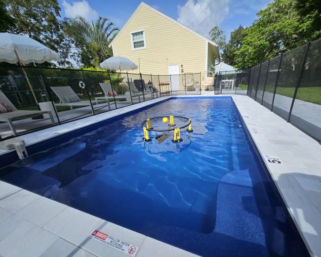 The image shows a rectangular swimming pool with a net setup for a game, surrounded by lounge chairs and umbrellas in a residential backyard.