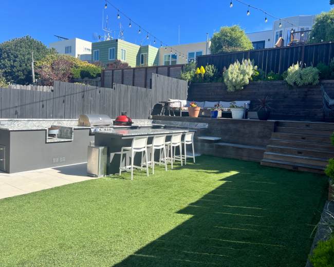 The image shows an outdoor kitchen area featuring a barbecue grill, seating, and a lawn with steps leading to a higher level adorned with various plants.
