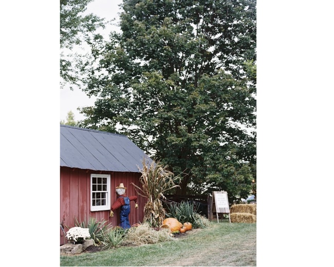 A person in a straw hat leans against a red barn surrounded by pumpkins and autumn decorations.