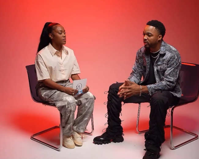 A man and a woman are seated in modern chairs, facing each other against a red backdrop during an interview.