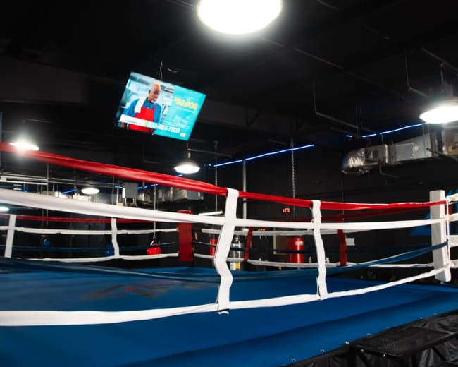 The image shows an empty boxing ring with blue flooring and colored ropes inside a gym, illuminated by overhead lights and featuring a wall-mounted screen.
