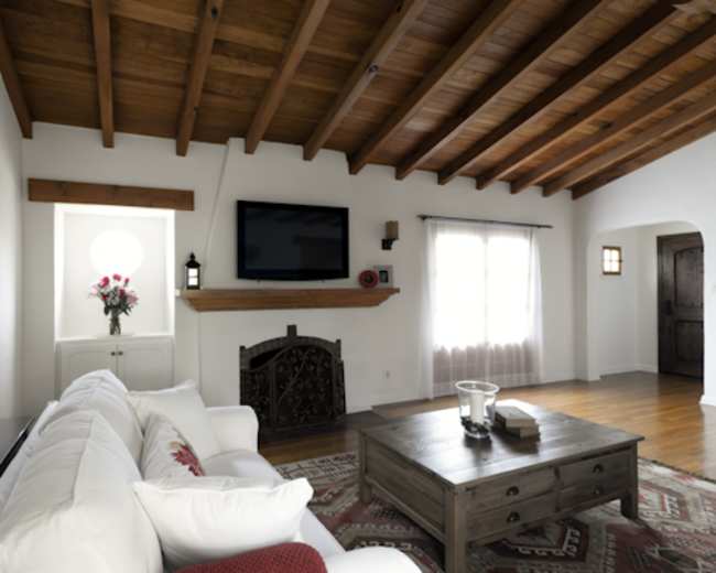 The image shows a living room with a white sofa, a wooden coffee table, a flat-screen TV mounted above a wooden mantel, and large windows with sheer curtains.