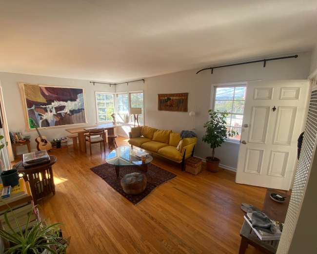 A spacious living room features a yellow sofa, a glass coffee table, a dining area with a wooden table, and large windows letting in natural light.