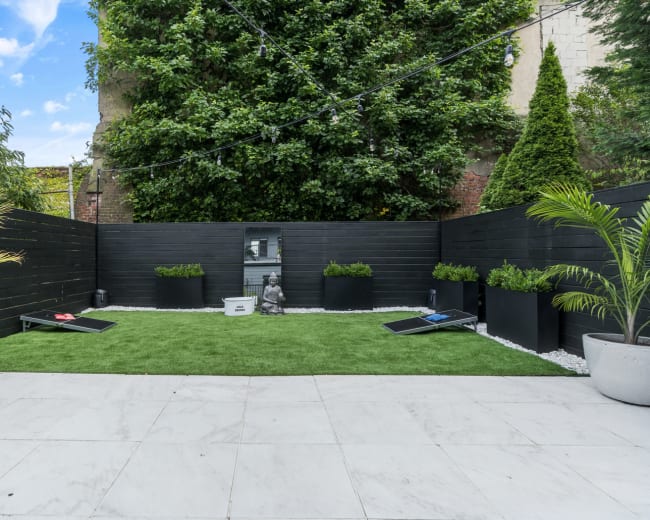 The image shows a small, enclosed garden with artificial grass, two lounge chairs, and large planters filled with greenery against a backdrop of stone walls.