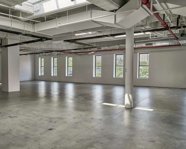 The image shows an empty, open commercial space with concrete flooring and multiple windows allowing natural light.