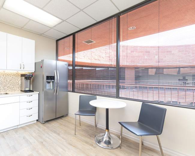 A modern kitchen area features a silver refrigerator, a sink, white cabinetry, and a small round table with two chairs beside large windows.