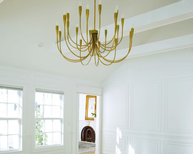 A spacious room features a gold chandelier, large windows casting light on herringbone wooden flooring, with an open doorway leading to another area.