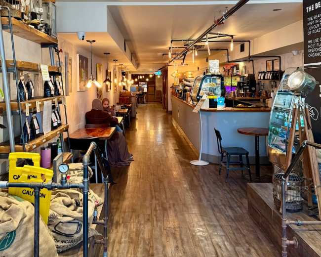 The image shows a cozy café interior with wooden flooring, shelves displaying various products, and a service counter at the end.
