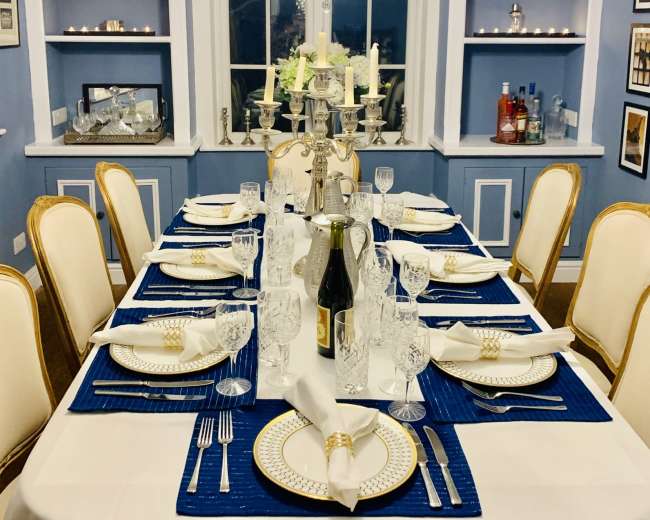 A formal dining table is set with elegant dinnerware and glassware beneath a chandeliers in a blue-walled room.