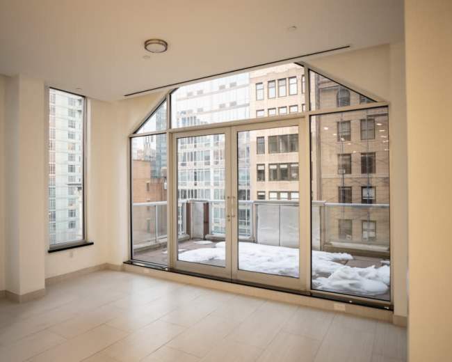 The image shows a vacant room with large windows overlooking a snow-covered balcony and buildings in the background.