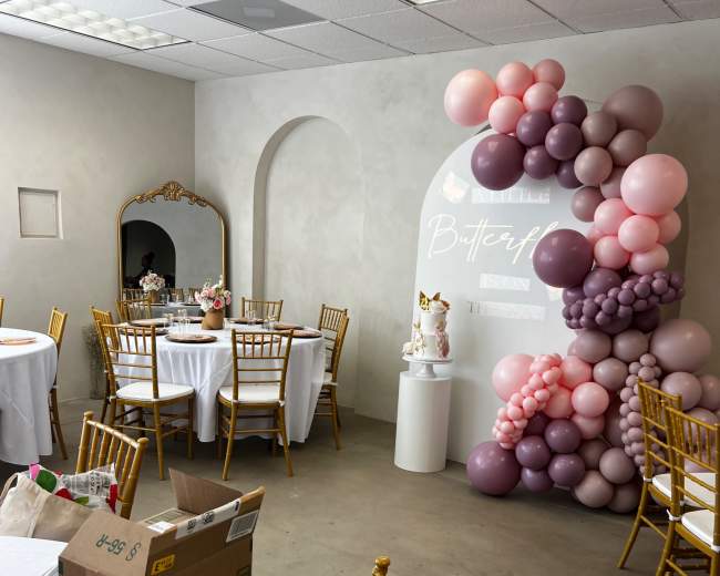 The room features two round tables with white tablecloths and gold chairs, decorated with balloons in shades of pink and purple along one wall, and a mirror reflecting the decor.
