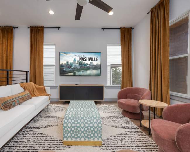 A modern living room with a white sofa, a patterned blue coffee table, two pink chairs, and a large wall-mounted TV displaying "Welcome to Nashville."