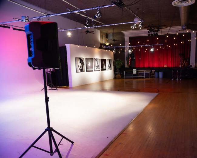 The image shows a spacious indoor venue with a stage at the far end, a backdrop of lights, and a display of black-and-white photographs on the left wall.