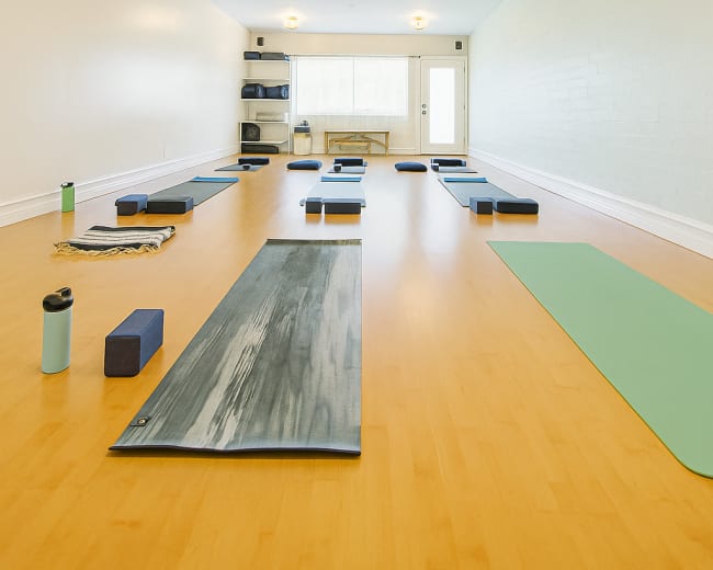 The image shows a spacious, well-lit yoga studio with several yoga mats arranged neatly on a wooden floor, and props like blocks and water bottles placed nearby.