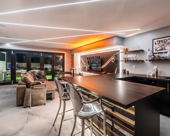 The image shows a modern living space with a bar area, featuring a sleek countertop and stylish seating, alongside a couch and a wall-mounted television.