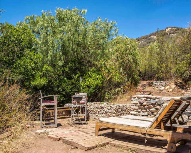 A wooden lounge chair and a stone barbecue area are set against a backdrop of greenery and hills.