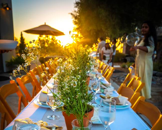 A long table set for an outdoor dinner features flower arrangements and glasses of water, with a sunset in the background.