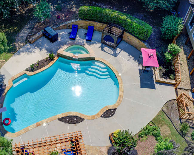 The image shows an aerial view of a curved swimming pool surrounded by patio furniture and landscaped greenery.