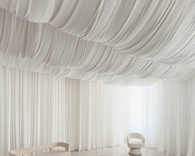 The image shows a minimalist room with draped white fabric covering the ceiling and walls, a smooth concrete floor, and simple furniture including a bench and a chair.