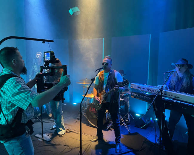A band performs on stage with a cameraman recording them in a dimly lit setting with colored lighting and smoke.