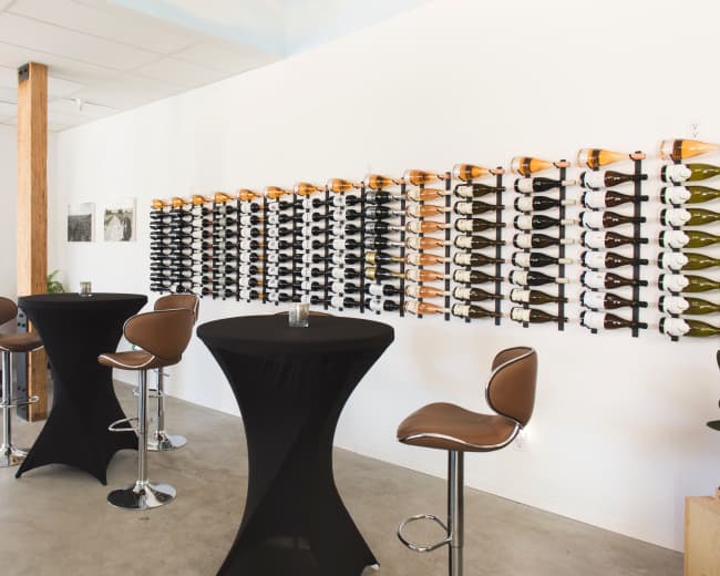 A modern interior of a wine bar with a wall-mounted wine rack displaying various bottles and several high-top tables with black tablecloths.