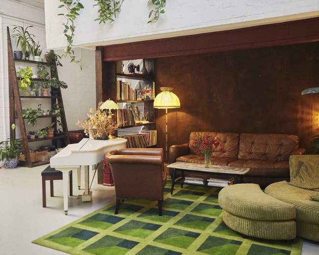 The image depicts a modern living space with a brown leather sofa, a green patterned rug, a warm lamp, and potted plants lining the shelves and walls.