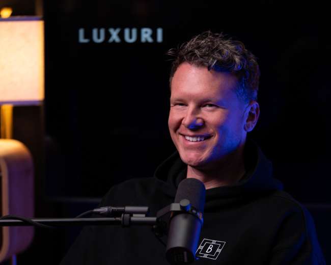 A man with curly hair is sitting in a chair, wearing a black hoodie and smiling while holding a microphone in a modern studio setting.