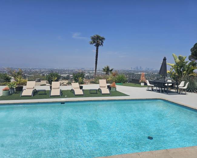 A clear swimming pool with lounge chairs and a dining table overlooks a city skyline surrounded by greenery.