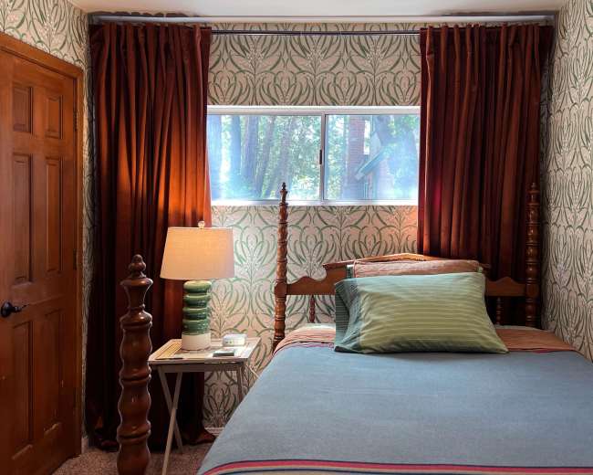 A small bedroom features a single bed with a green and striped blanket, a bedside lamp on a table, and patterned wallpaper with a window showing greenery outside.