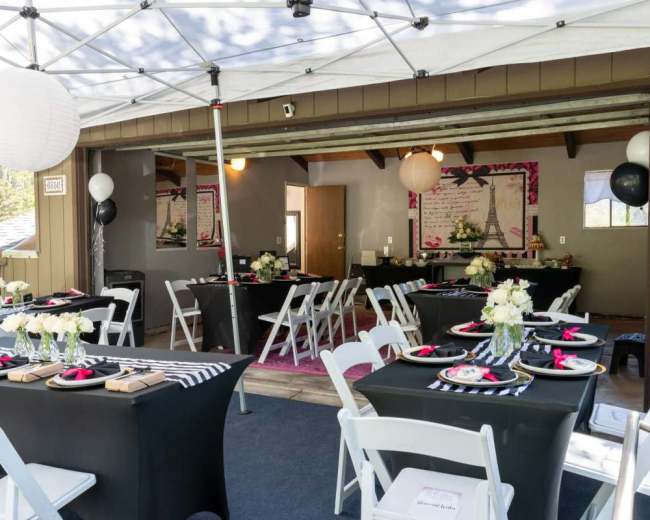 A covered outdoor space features several dining tables set up with black tablecloths, decorative plates, and floral centerpieces.