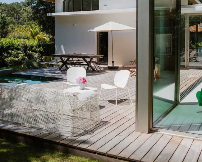 The image shows a modern house with a wooden deck, outdoor seating, and a swimming pool surrounded by greenery.